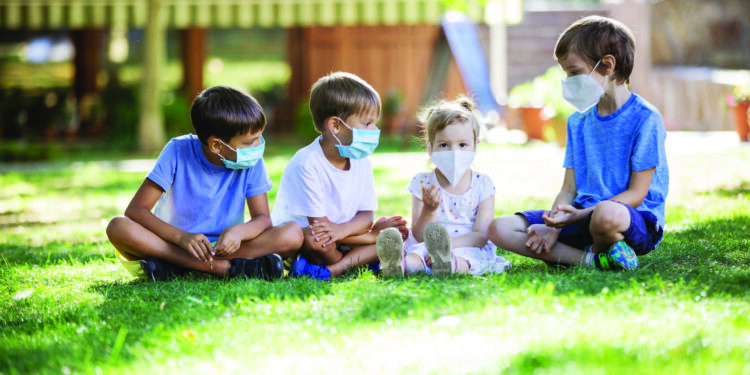 Young children in protective masks on faces outdoors. Quarantine. Kids wearing safety masks while sitting on grass in park. Coronavirus prevention.