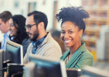 Pretty African American woman with natural curly hair smiling at camera while using computer in library or lab, to continue college education or job training. Other diverse adults can be seen in backgroud, also using computers.